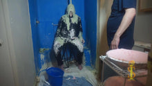 Man sitting on stool has green slime fall on his face and head as he looks up into it. The slime splatters onto shirt, pants, and sleeve
