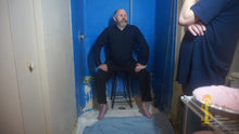 Man with receding hair and gray and black beard sits on stool thinking. 