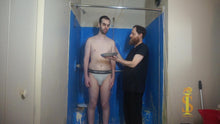 Two men. One wearing only briefs, other man in all black. The man in black holds a pie in front of the man in briefs. The man in briefs looks at the pie cautiously