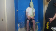 Two men, one in a white shirt and black shorts. Green slime is falling on him and covering his face. Other man, wearing black, stands  off to the side, looking at him