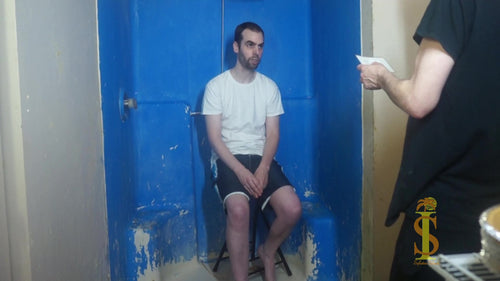 Two men, one sitting on stool in white shirt and black shorts, looking at other man wearing all black. The man wearing all black holds an index card